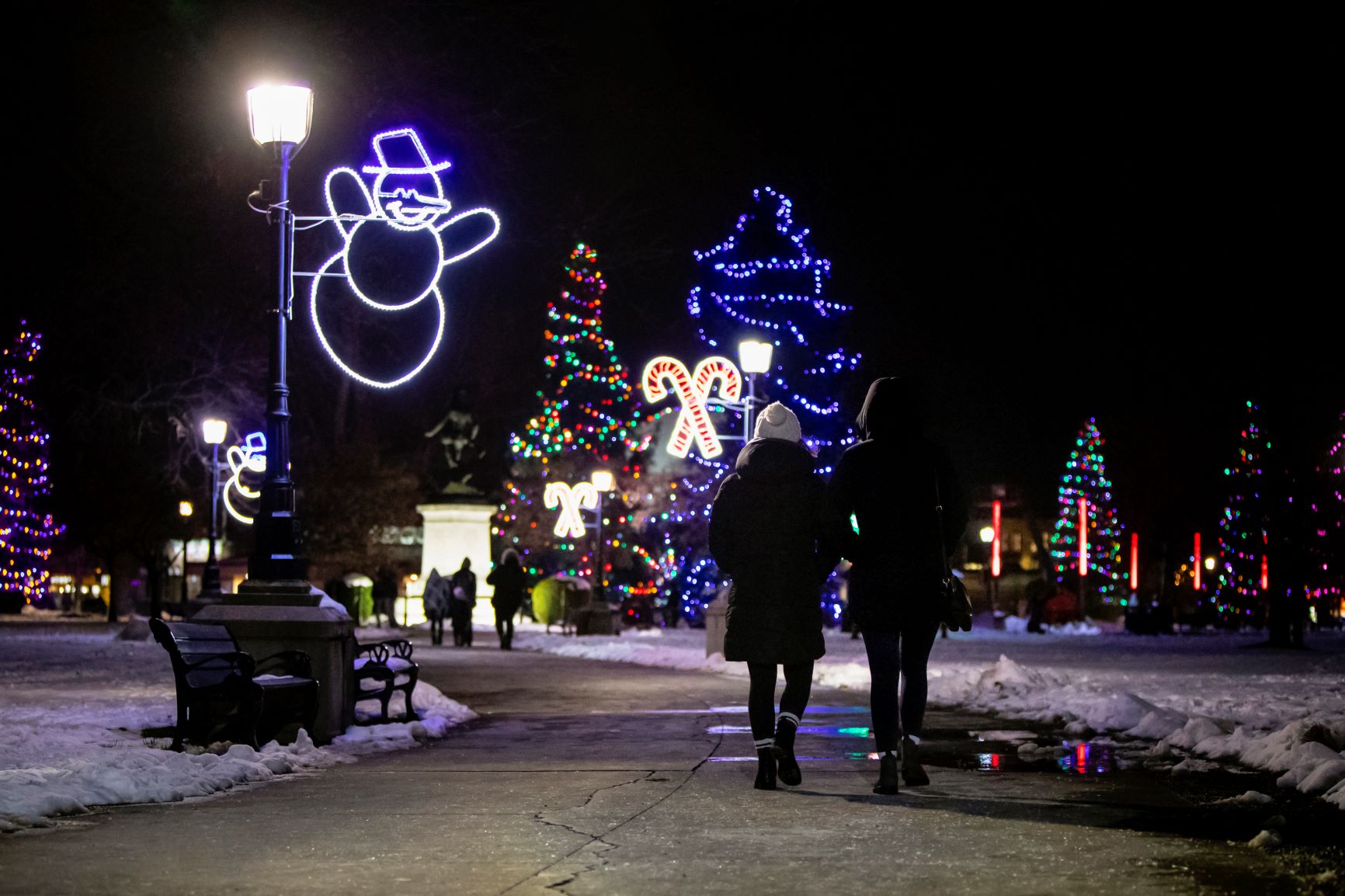 Two people walking through park with holiday lights and decorations along the parkway