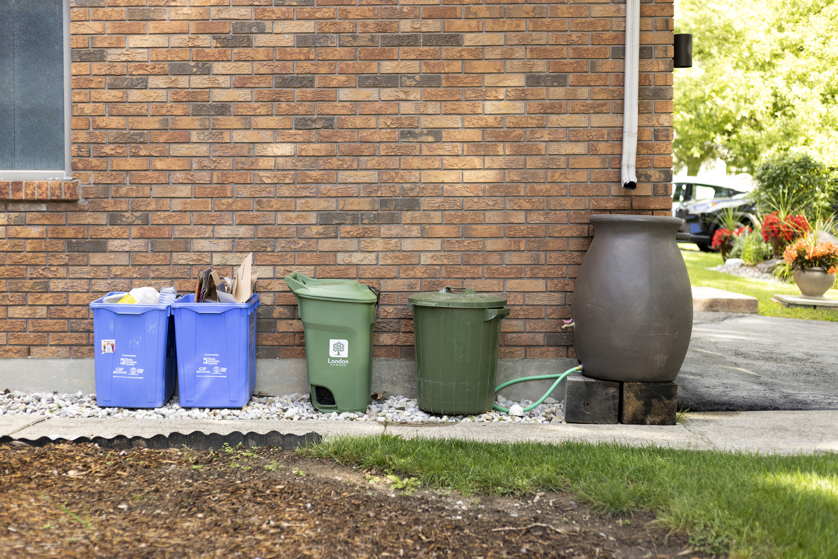 Two blue boxes, a Green Bin, a garbage container, and a rain barrel up against the exterior of a home's garage. 