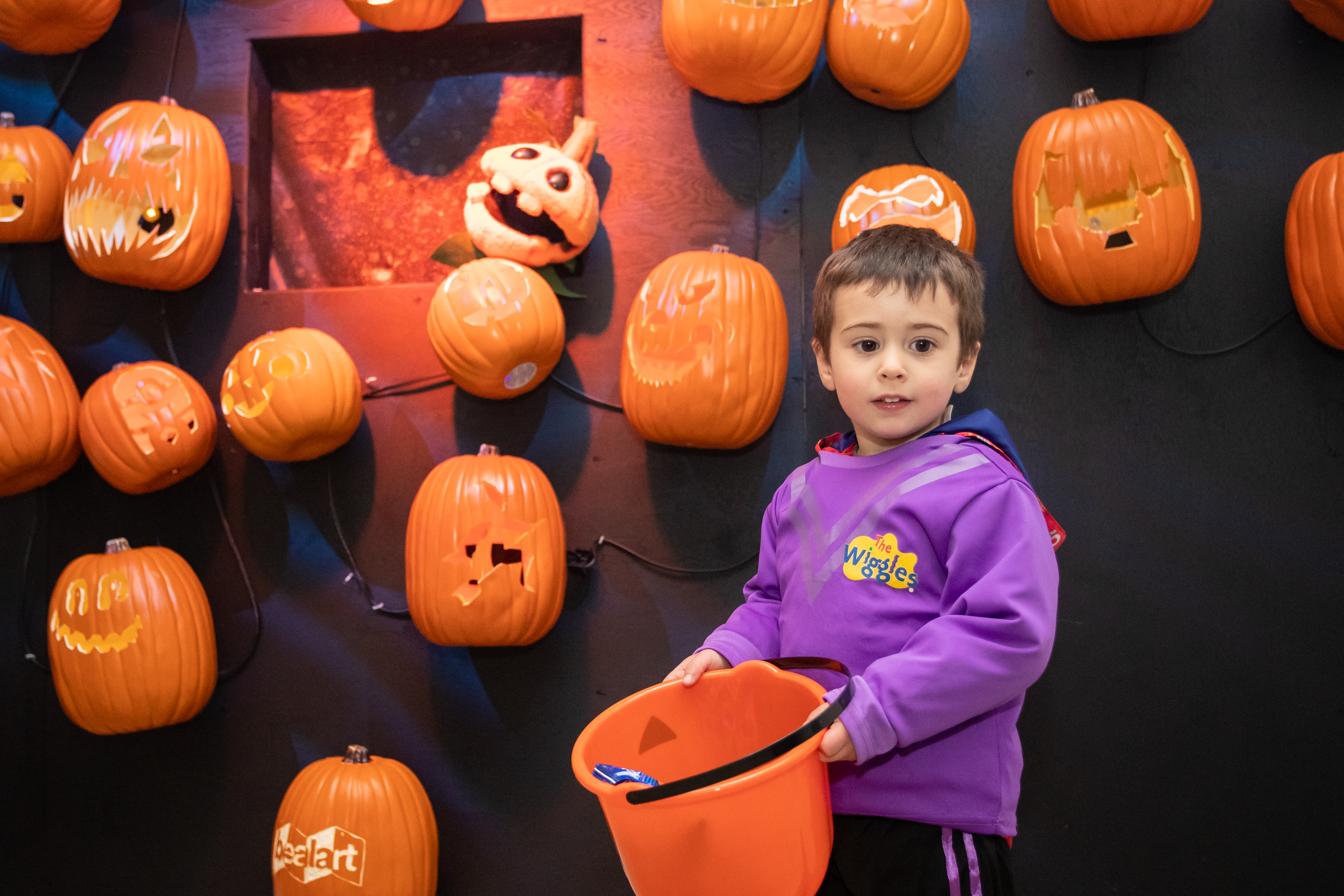 Child with a Wiggles custome holding a trick-or-treat basket and smiling at the camera. Halloween decorations, including pupkins, are seen in the backgournd. 