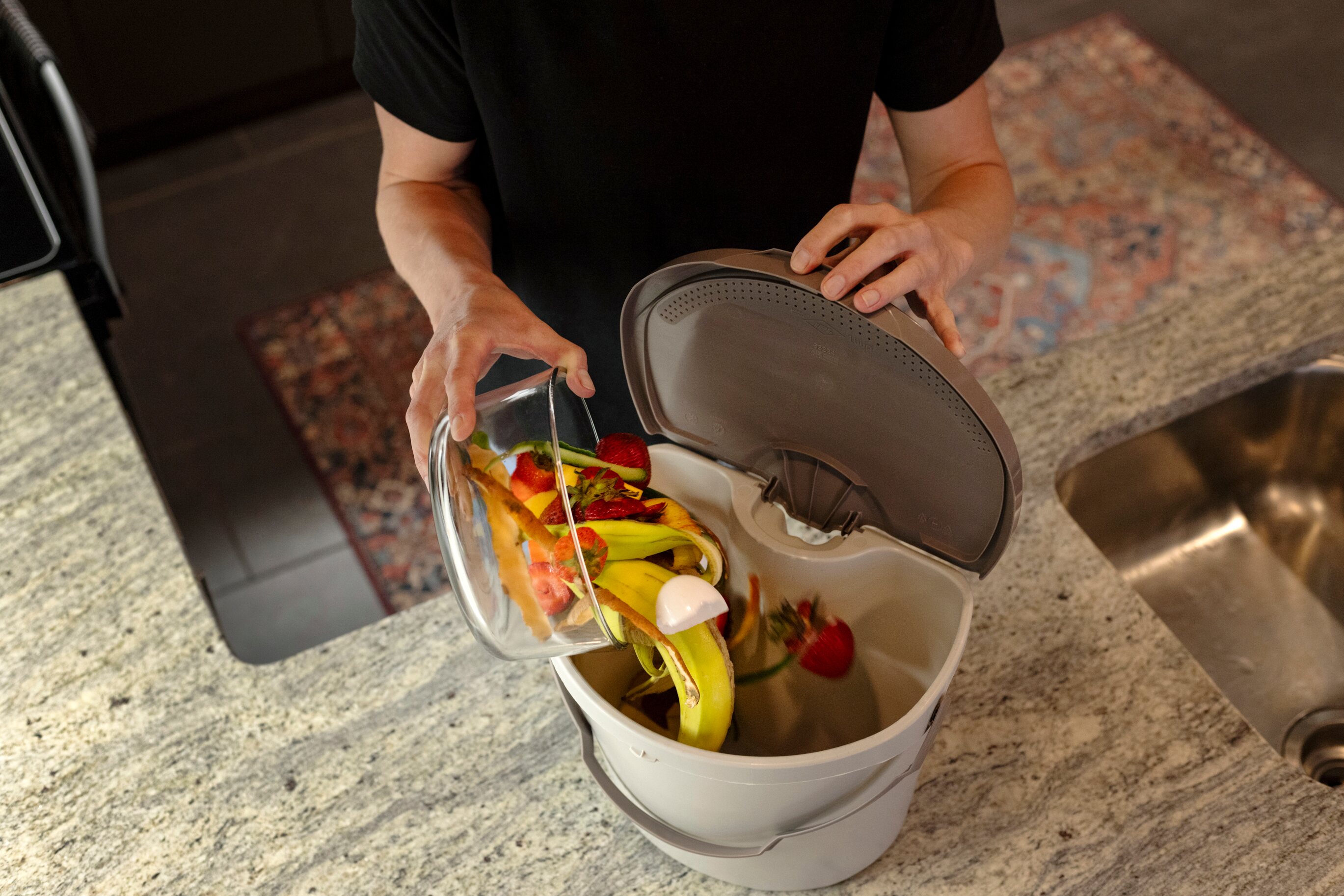 A woman pours food scraps from a glass jar into a Kitchen Container while standing in their kitchen. 