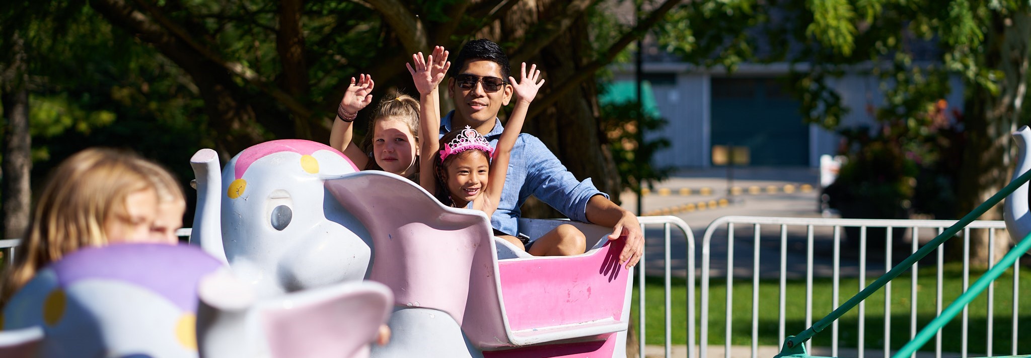 An adult and two children ride in the pink and white elephant seat on the mechanical elephant ride at Storybook Gardens. The two children have their hands in the air.