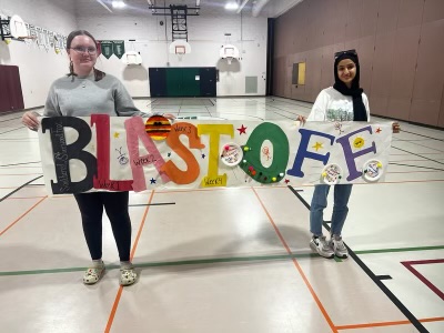 Two youth hold a colourful, handpainted sign that says "Blast off" in a gym during the summer day camp program.