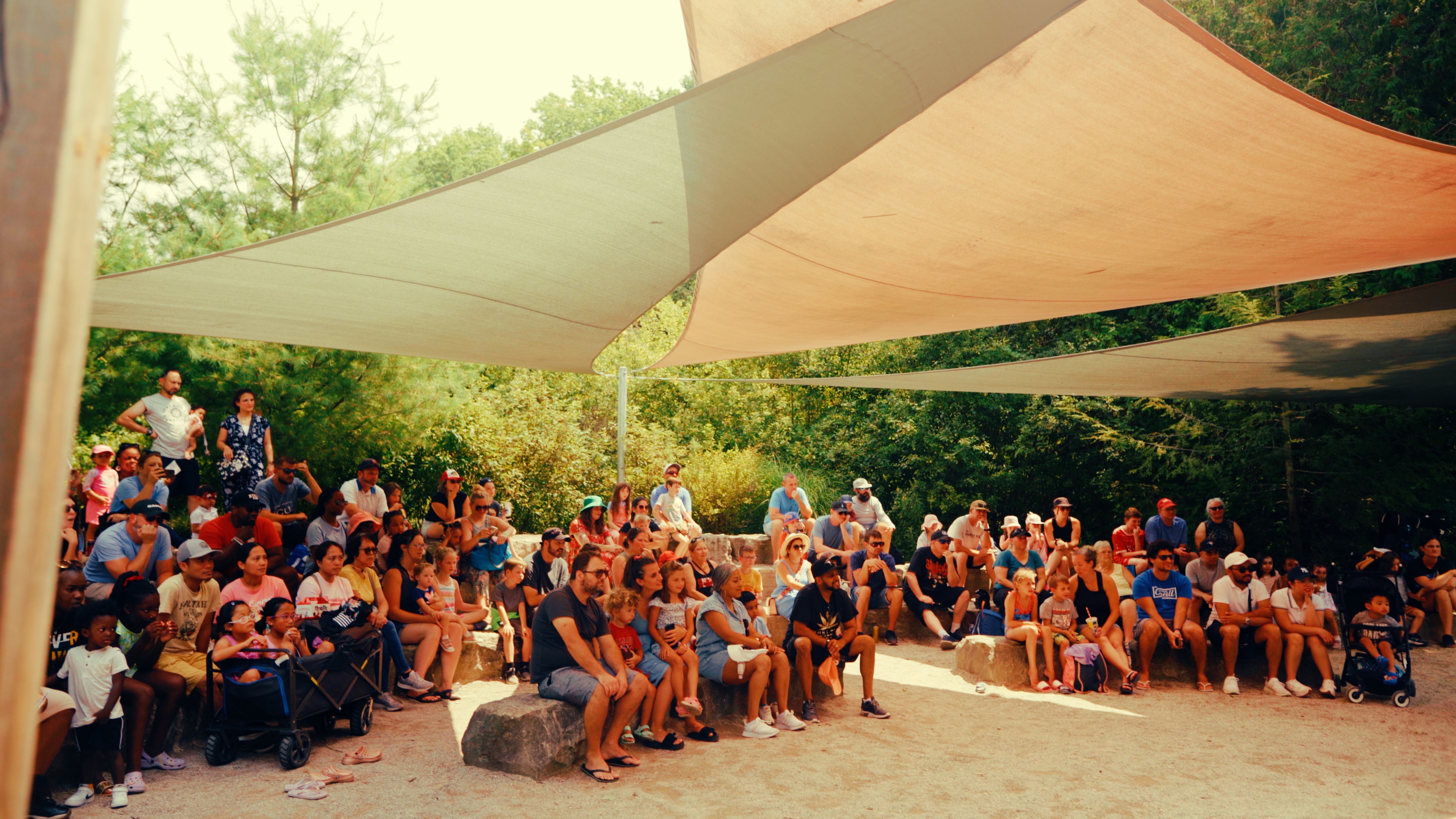 A large group of adults and children sit underneath a tent to watch a performance at Storybook Gardens in the summer.