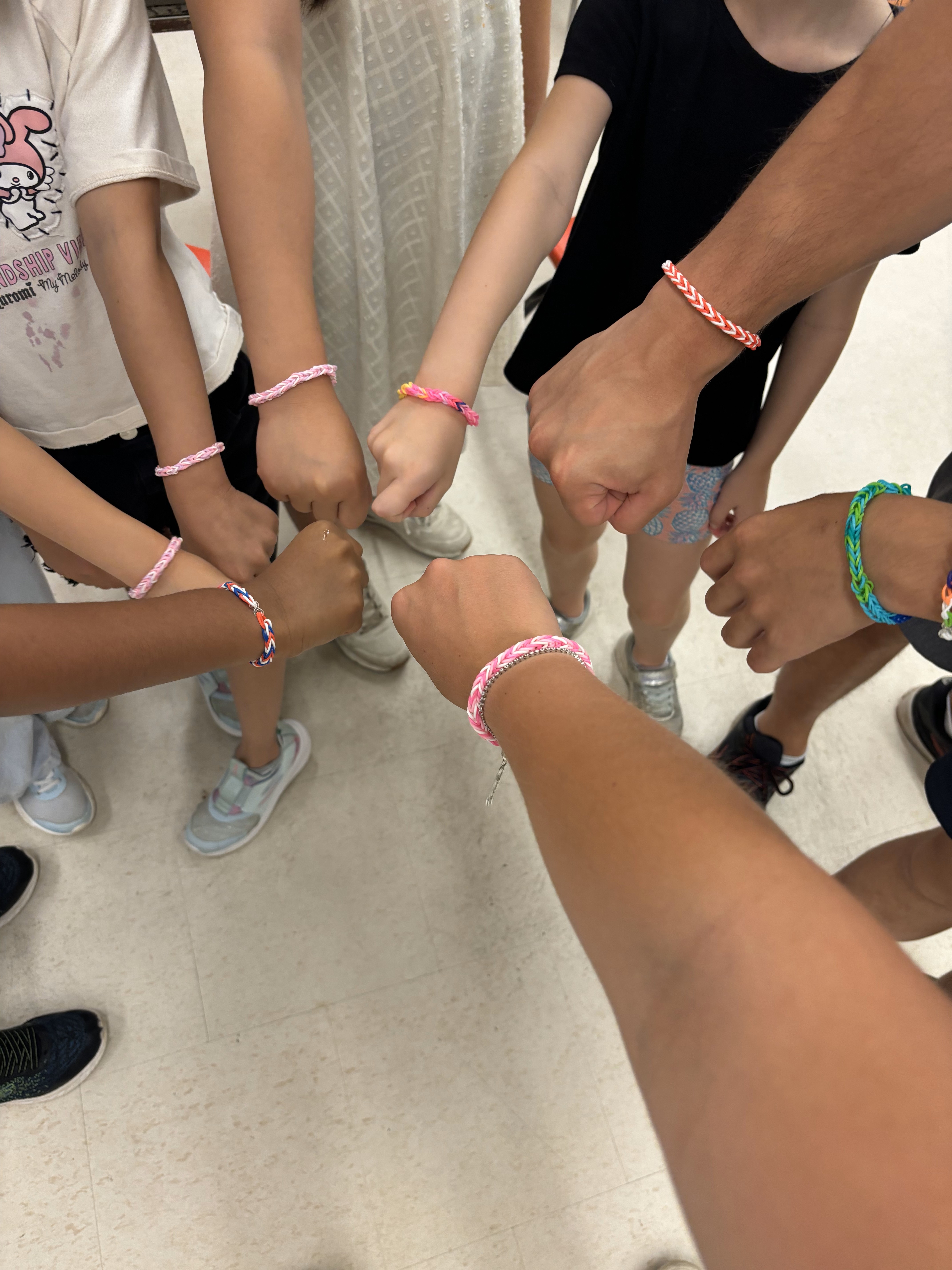 Eight different hands with bracelets on their wrists are aimed in a circle with hands making a fist. 