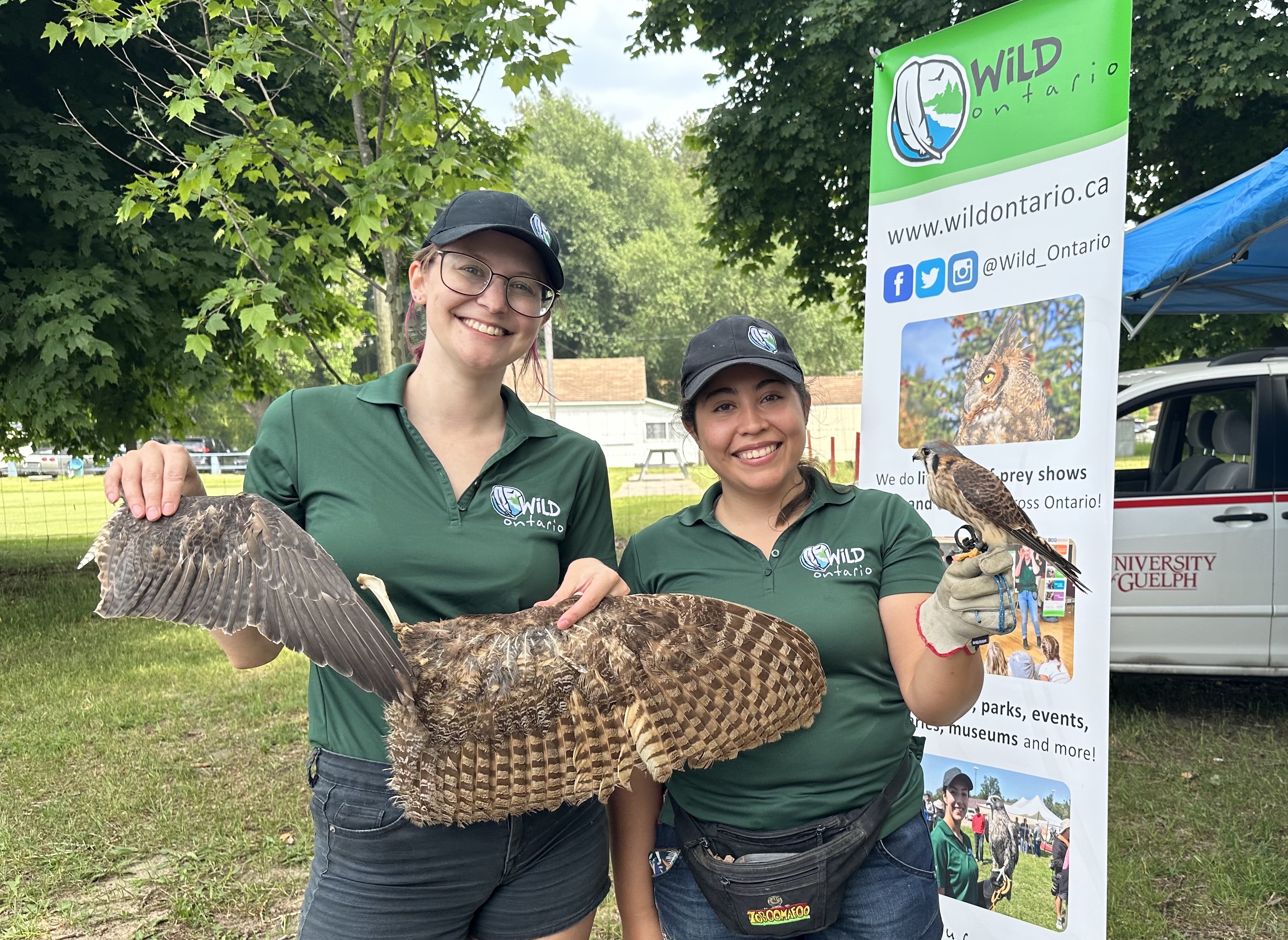 Two people wearing green "Wild Ontario" shirts stand outdoors holding birds of prey with outstretched wings. A banner for Wild Ontario with their website and social media handle is visible behind them. Trees and a building are in the background.
