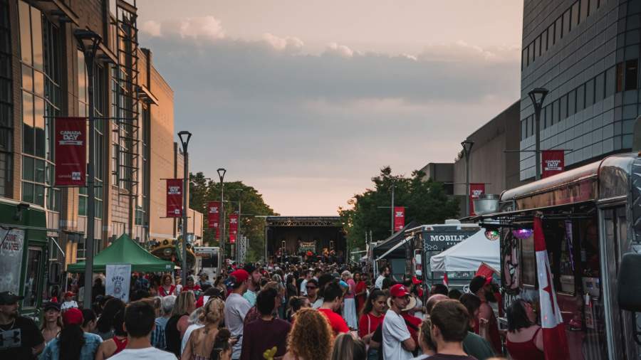 A crowd of people on Dundas Place for the Canada Day celebration. There is a stage in the far distance where musicians will perform. A sunset can be seen. 